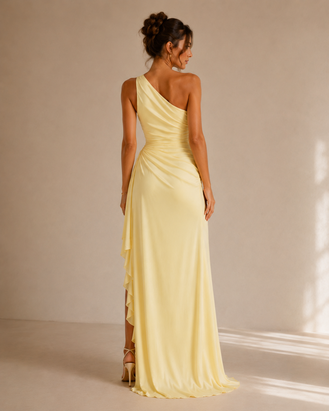 A woman stands with her back to the camera wearing a pale yellow one-shoulder floor-length gown in a softly lit studio with a warm beige wall and floor.