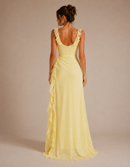 A woman modeling a pale yellow sleeveless maxi dress from the back, featuring ruffled shoulder straps, a deep V-neckline, and cascading ruffles down the side, set against a solid beige studio background.