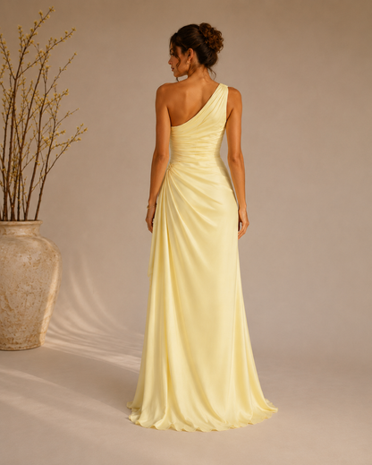 A woman stands with her back to the camera wearing a floor-length one-shoulder satin gown in a pale buttery yellow. The dress features a draped, gathered bodice and a flowing skirt.