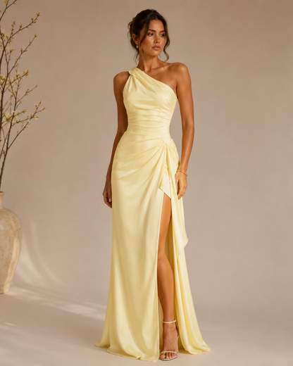 A woman models a yellow, one-shoulder satin maxi dress with a high thigh slit and gathered ruching at the waist, set against a warm beige studio background with a decorative vase on the left.