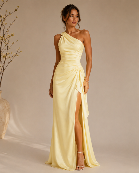 A woman models a yellow, one-shoulder satin maxi dress with a high thigh slit and gathered ruching at the waist, set against a warm beige studio background with a decorative vase on the left.