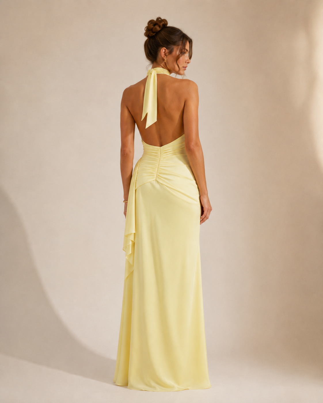 A woman modeling a pale yellow halter-neck maxi dress from the back, featuring a draped open back and gathered waist, set against a warm beige studio background.